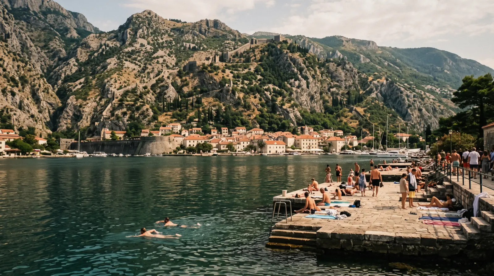 Swimmers off the stone slabs of the Kotor waterfront with the bay and Mount Lovcen behind